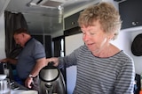 A woman pours a cup of tea while a man makes a coffee. They are both inside a caravan