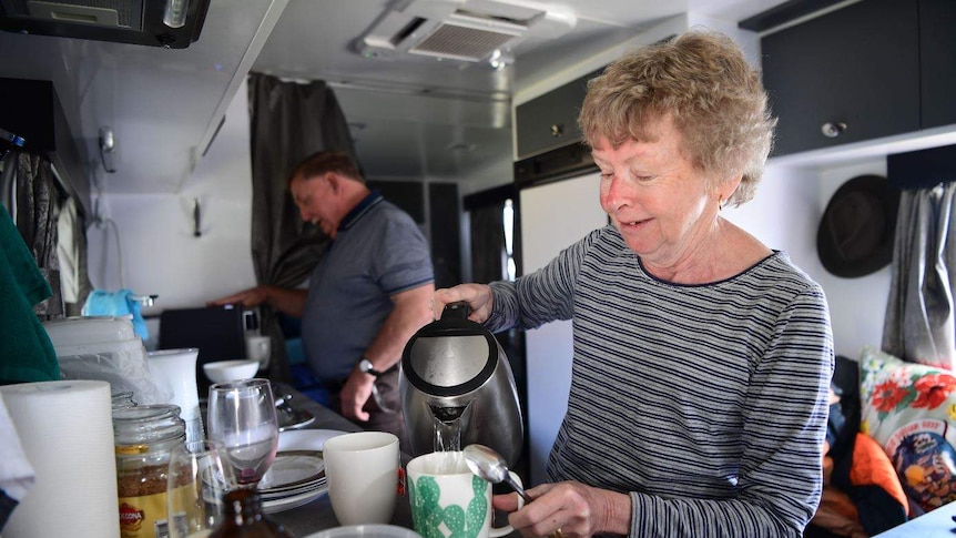 A woman pours a cup of tea while a man makes a coffee. They are both inside a caravan