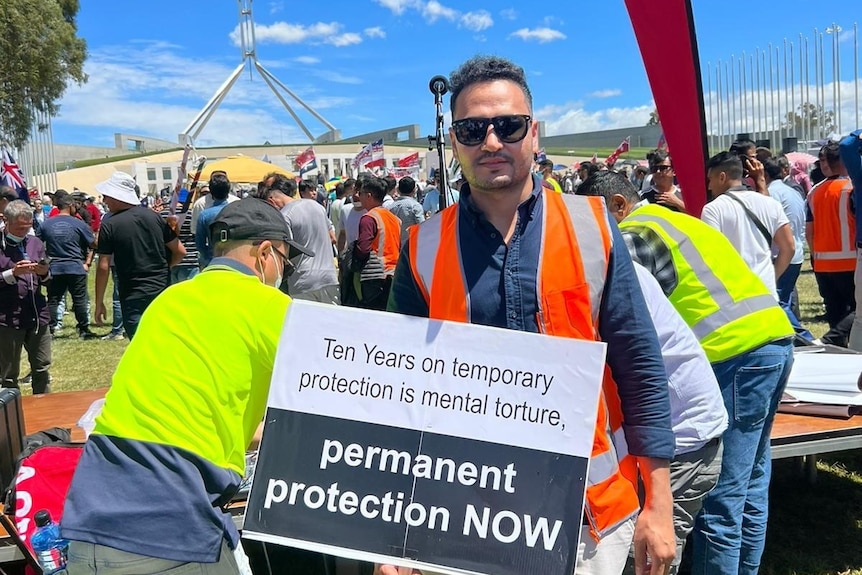 A man in a high-vis vest stands in a crowd holding a sign that says 'permanent protection now'