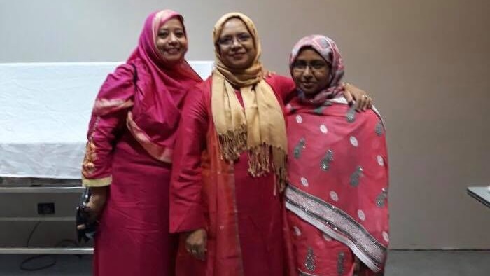 Three women smile at the camera. They are wearing colourful pink and red dresses and headscarves.