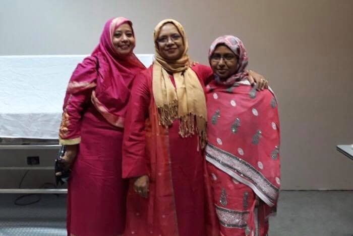 Three women smile at the camera. They are wearing colourful pink and red dresses and headscarves.