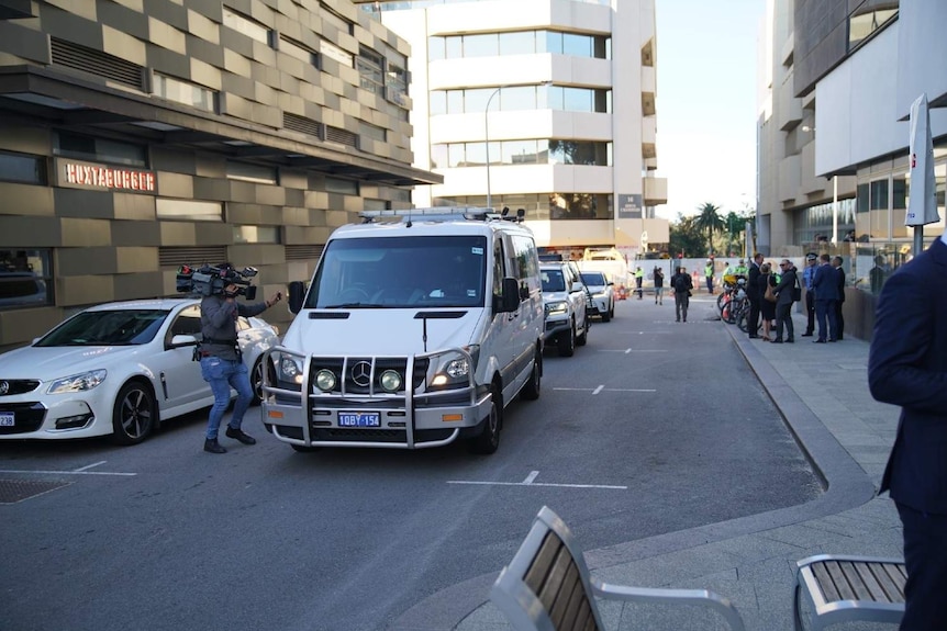 A white prison van on a city street with a large crowd waiting on the side of the road.