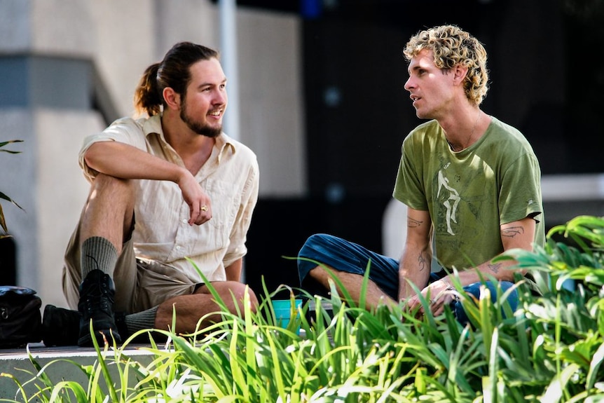 two young men one with a beard and goatee sitting on the grass talking to each other