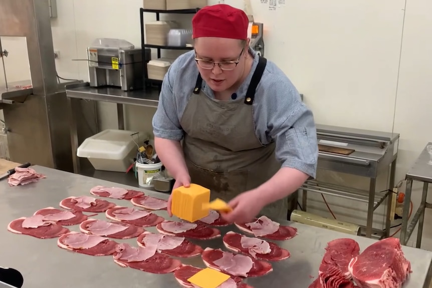 A woman in a butcher's outfit lays cheese on pieces of steak and ham laid out flat