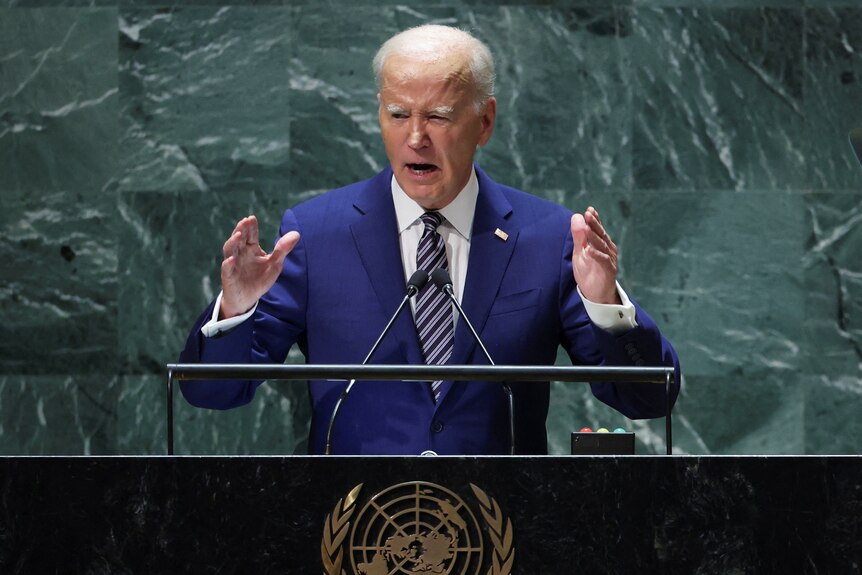 Joe Biden gestures with his hands while standing at a podium and speaking. He wears a blue suit and striped tie.