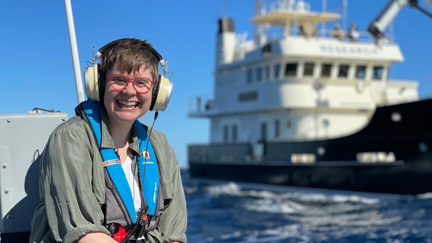 Dr Ann Jones wears large headphones and sits at the edge of the water with a large ship in the background.