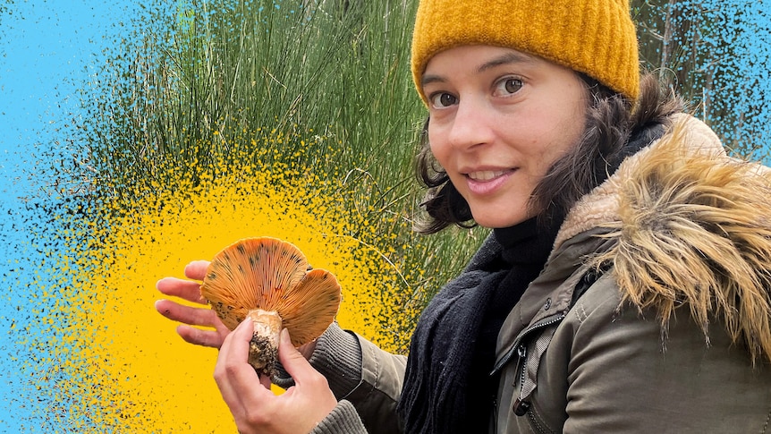 Zoe, a young woman wearing a coat and yellow beanie, looks over her shoulder holding a large orange mushroom