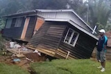 A man stands next to a tilted and damaged house.