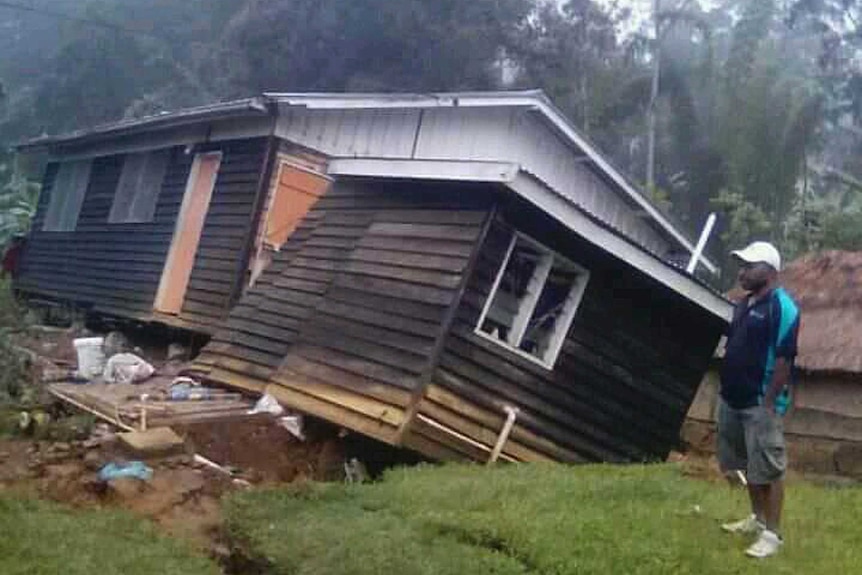 A man stands next to a tilted and damaged house.