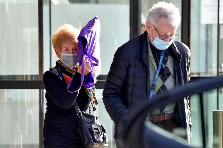 Two elderly people wearing face masks and jackets, with a woman holding a purple bag in front of her head