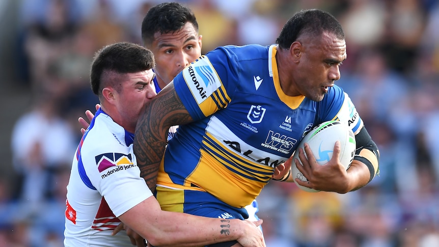 A Parramatta Eels NRL player holds the ball with his right hand as he is tackled by two Newcastle Knights opponent.