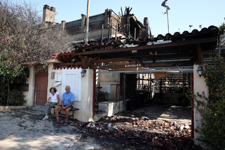 A couple sit on a bench outside a house which has been damaged by fire. 