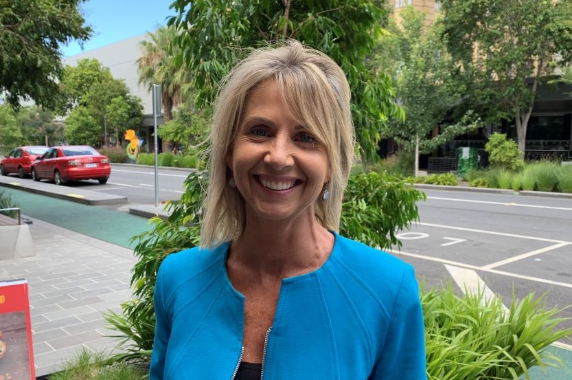 A blonde woman in a light blue blazer stands on a tree-lined street beside a bike lane and smiles.