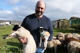 A Farmer holds two lambs, one normal size and the other very small, wearing a woolen jacket