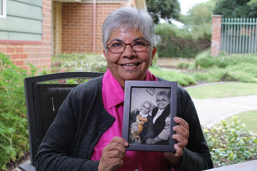 Deidre de Souza sits on a chair outside smiling and holding a black and white photo of her with her mum.