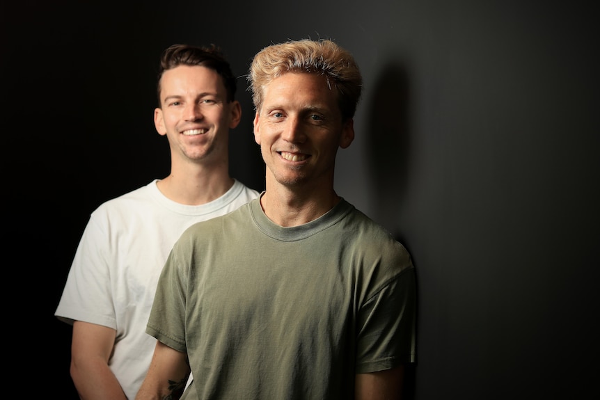 Sam wears a white t-shirt and Brett wears a green t-shirt, both smiling with a black wall behind them.