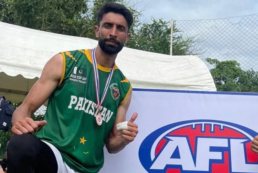 A man in a green singlet wears a medal and gives a thumbs up, standing next to a trophy and an AFL Asia sign