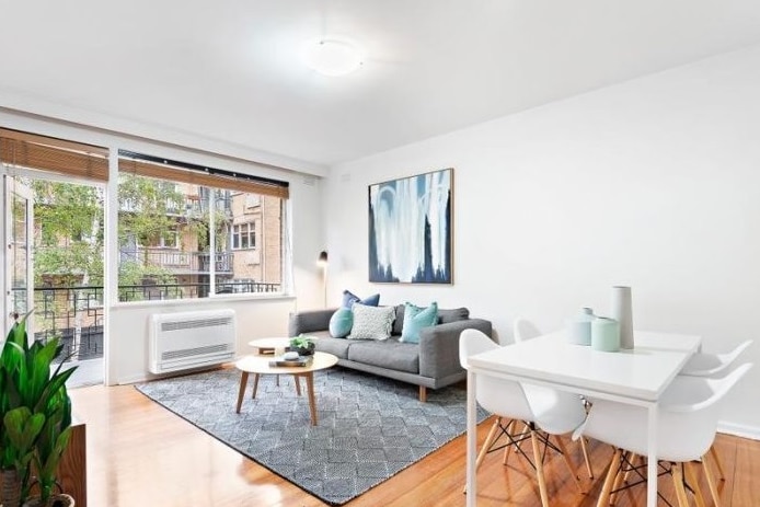 An apartment, with white walls and wooden floorboards, contains furniture including a white dining setting and a blue couch.