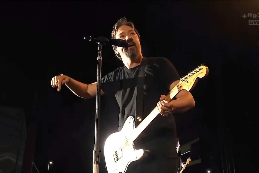 A band's frontman in a black shirt holding a guitar and pointing out towards the crowd.