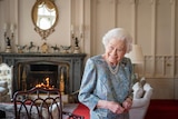 Britain's Queen Elizabeth II smiles while standing in a formal drawing room with a fire and ornate furnishings. 