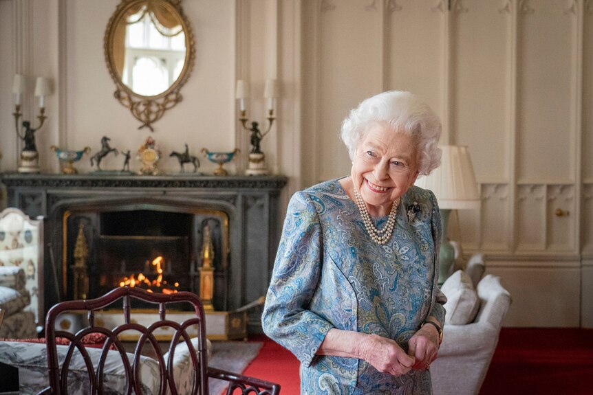 Britain's Queen Elizabeth II smiles while standing in a formal drawing room with a fire and ornate furnishings. 