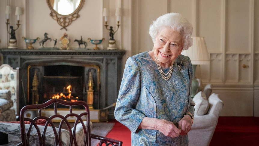 Britain's Queen Elizabeth II smiles while standing in a formal drawing room with a fire and ornate furnishings. 