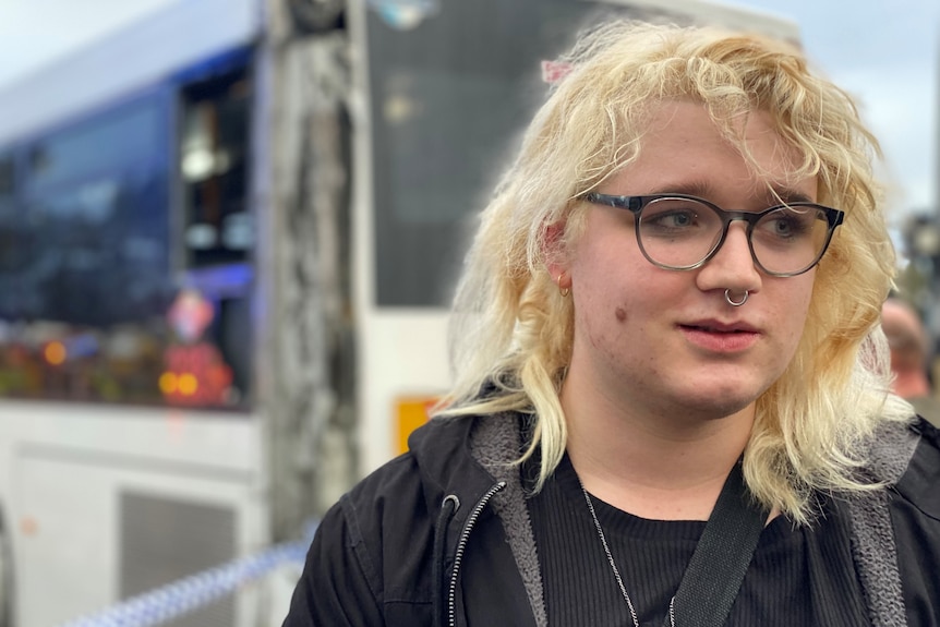 A blond-haired man with glasses and black top in front of a damaged bus