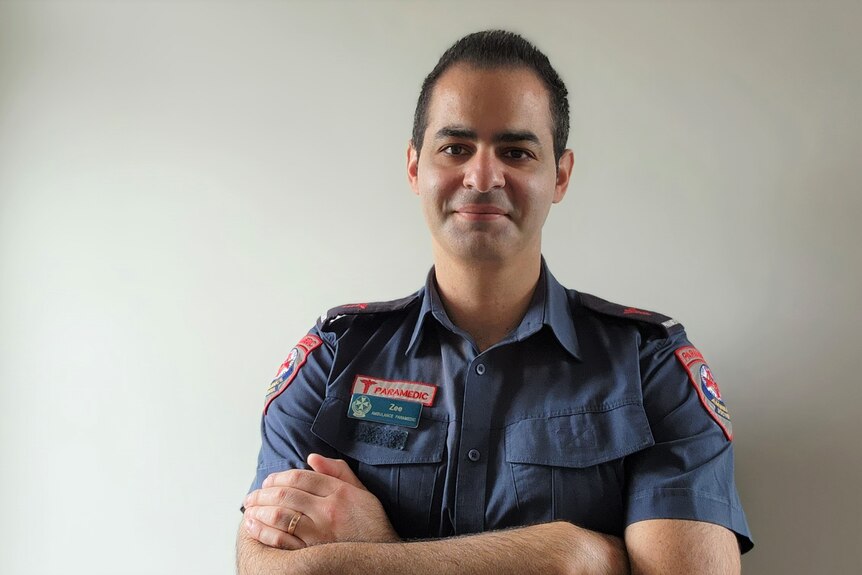 Head and shoulders profile of a smiling man in an paramedic uniform,k dark cropped hair, arms crossed, blank wall behind.