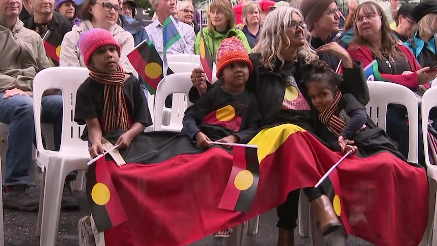 A woman and three children sitting on plastic chairs waving Aboriginal flags and with a large flag draped over their laps