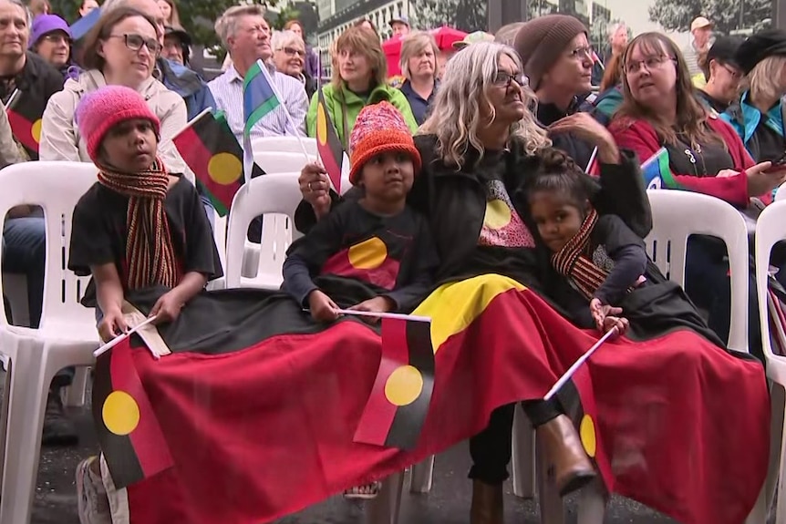 A woman and three children sitting on plastic chairs waving Aboriginal flags and with a large flag draped over their laps