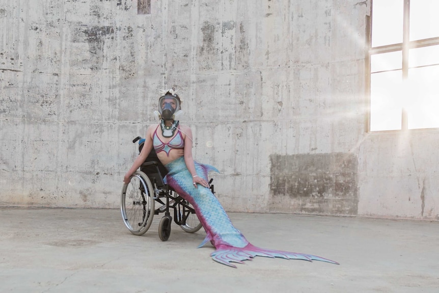 A woman in a mermaid costume and ventilator mask sitting in a wheelchair in a bright white empty warehouse
