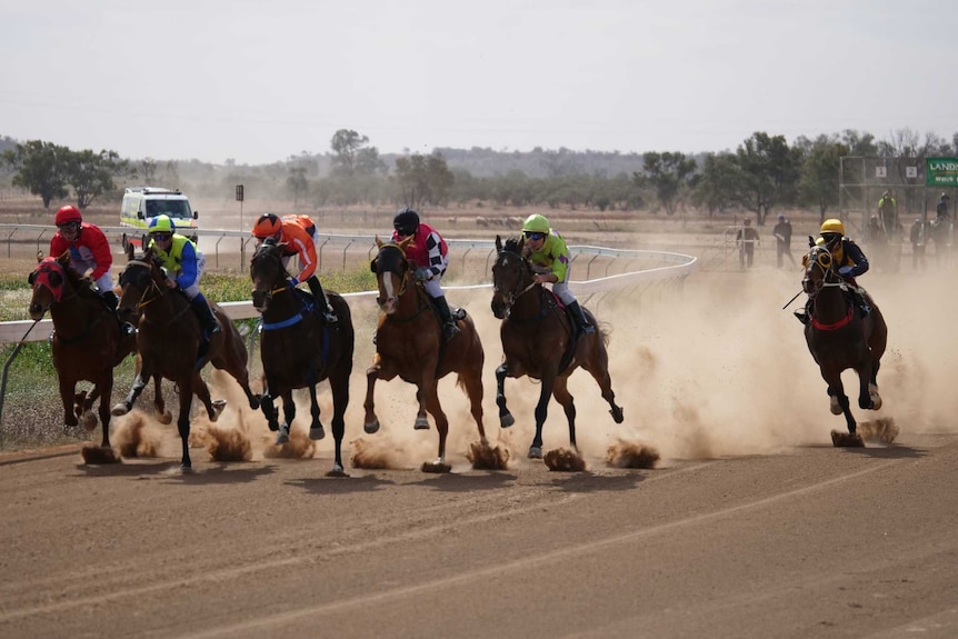 Six horses ridden by jockeys race along a very dusty track.