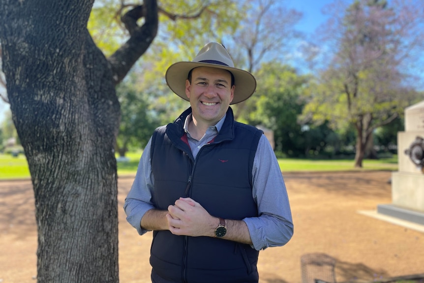 Sam Farraway smiles at the camera, wearing a wide-brimmed hat, standing outside under a tree.