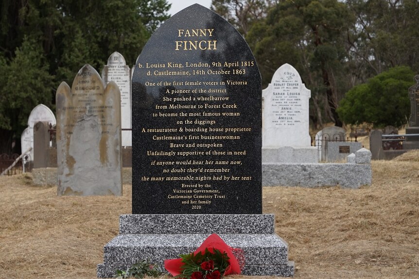 A bouquet of red roses lays at the foot of a black headstone with gold lettering on it