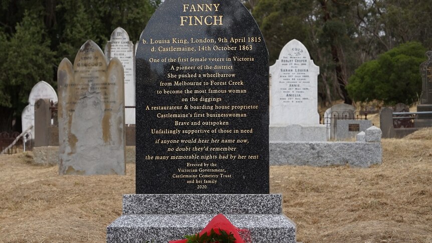 A bouquet of red roses lays at the foot of a black headstone with gold lettering on it