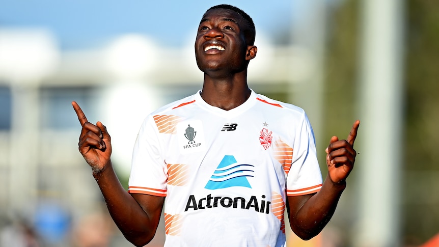 Cyrus Dehmie smiles and points to the sky wearing a white football shirt