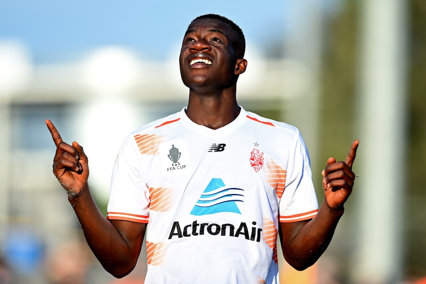 Cyrus Dehmie smiles and points to the sky wearing a white football shirt