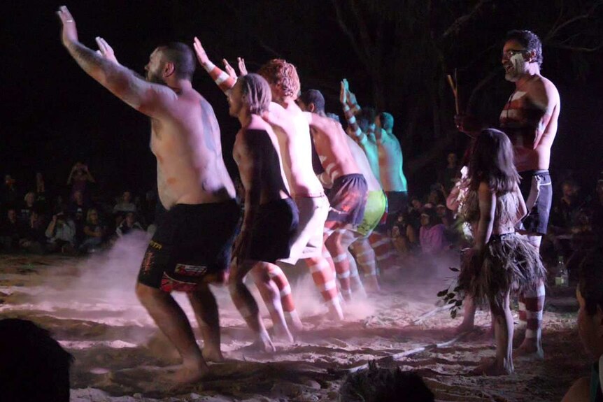 A line of Indigenous dancers hold their arms in the air and stamp their feet, raising dust.