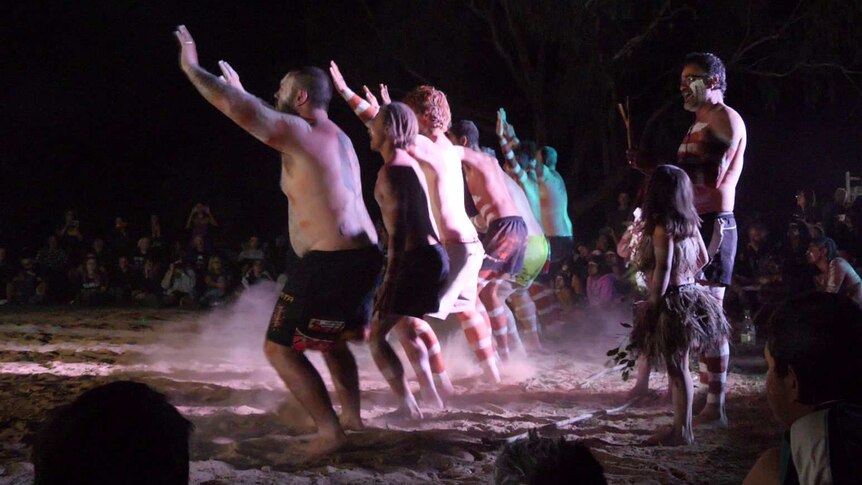 A line of Indigenous dancers hold their arms in the air and stamp their feet, raising dust.