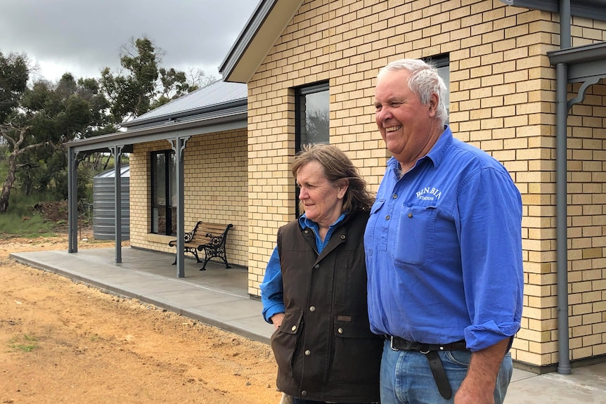 Arm in arm, a couple grin with their new home behind them