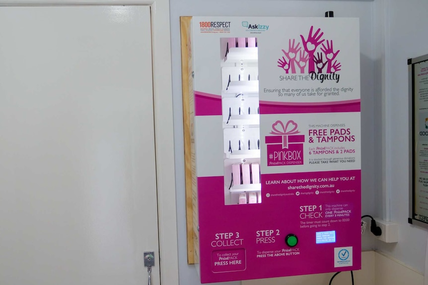 A photo of a small pink and white vending machine which is installed in a bathroom.