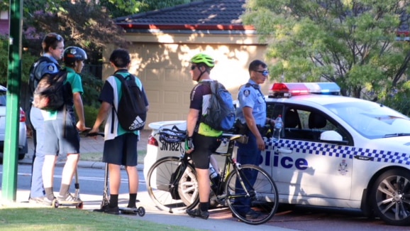 Two boys on scooters and a man with a bike talk to police on a street.
