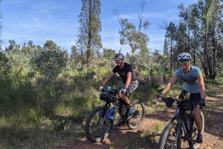 A man and a woman on their bikes on a dirt track surrounded by bushland.