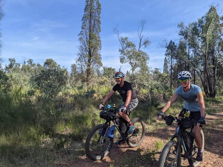 A man and a woman on their bikes on a dirt track surrounded by bushland.