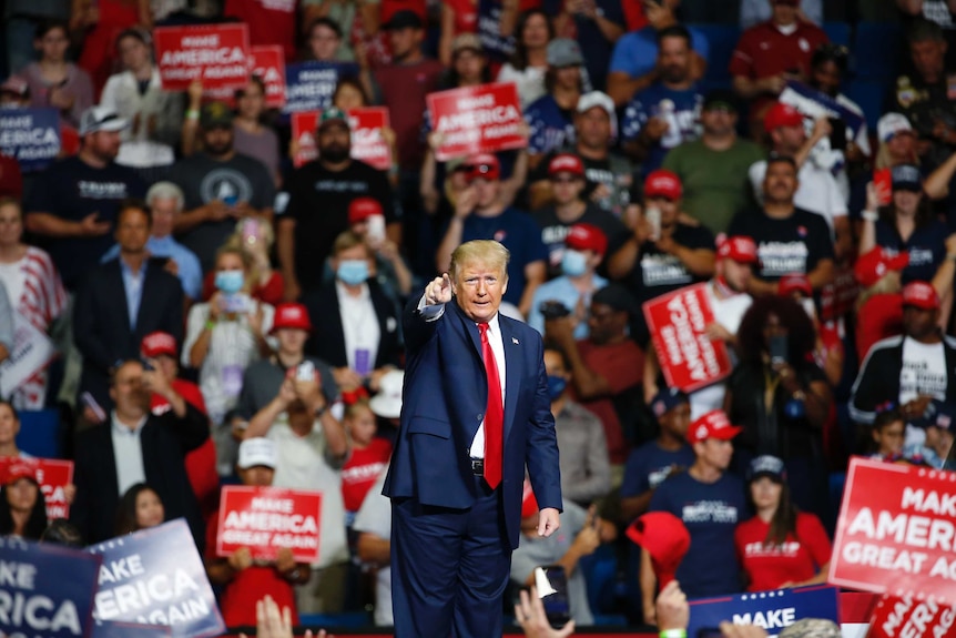 A man in a blue suit and red tie points as he speaks to a crowd of people holding placards.