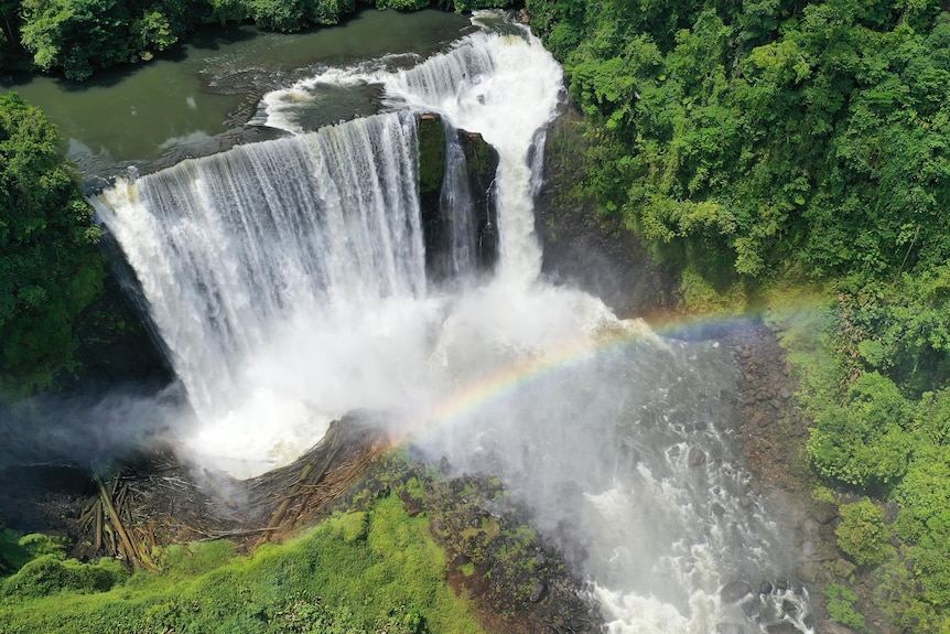 A large waterfall surrounded by green rainforest. A rainbow can be seen in the mist of the crashing water.