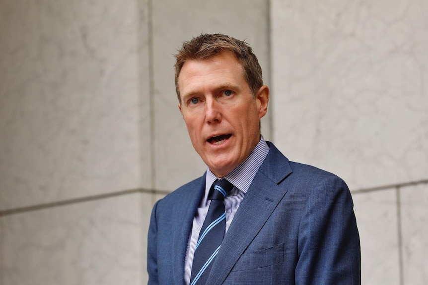 A man stands in front of a grey wall with his mouth open