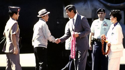 A man in a hat shakes hands with a man in a suit, holding a pink floral lei in his hands