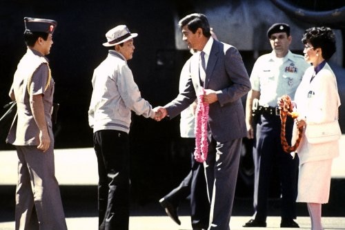 A man in a hat shakes hands with a man in a suit, holding a pink floral lei in his hands