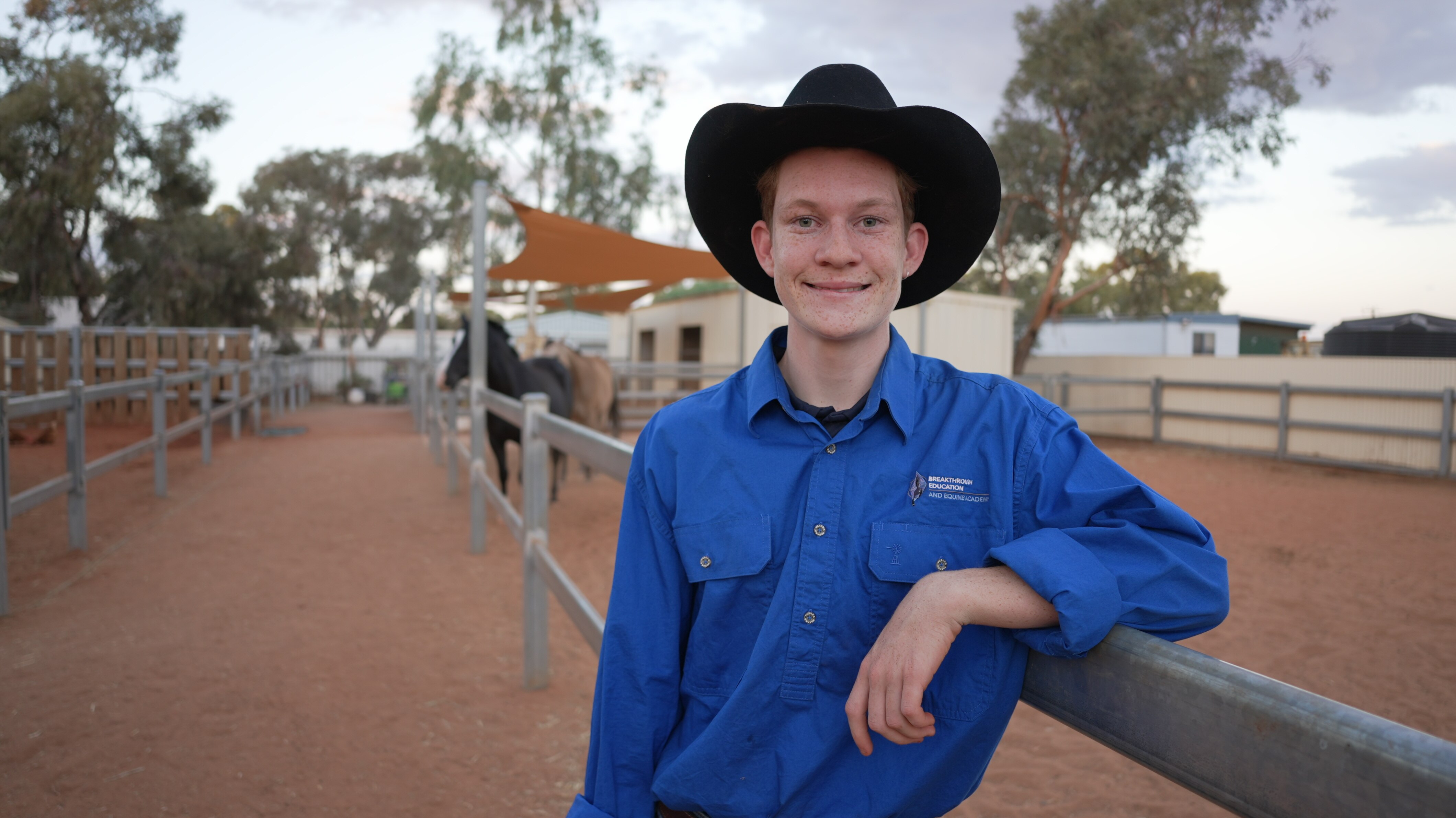 A teenage boy grins wearing an wide brim cowboy hat in front of horse stables. He leans on one of the fences. 